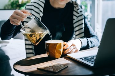 Close up of hand pouring a cup of medicine ball tea from a stylish transparent tea pot into a cup.