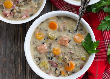 a white bowl of Creamy Ham and Wild Rice Soup on a wooden table with a spoon and fresh herbs