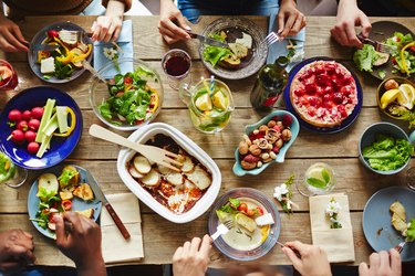 an overhead photo of a wooden table covered in serving bowls and platters of food with five people's hands visible enjoying their meals