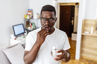 a man in his 40s wearing a white t-shirt taking a supplement at home holding a glass of water