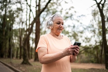 an older adult uses a mobile phone to play a workout playlist on silver headphone in a park as an example of workout motivation tips from fitness professionals