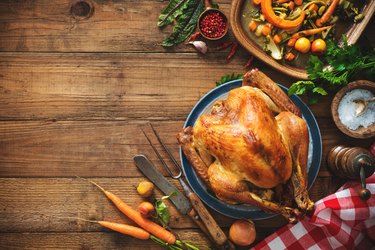 an overhead photo of a Thanksgiving turkey on a wooden table next to carrots, beets, greens and a red-checkered kitchen towel