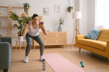 Person wearing white tank top and gray leggings exercising with dumbbells at home doing beginner CrossFit workout.