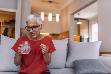 older woman wearing a red t-shirt and black glasses holding a multivitamin and a glass of water while sitting on a couch at home