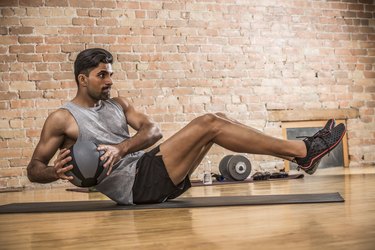 Person wearing gray tank top and black shorts doing Russian twist core exercise with medicine ball in gym.