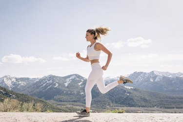 Young adult wearing white leggings and a white crop top running outside with mountains in the background