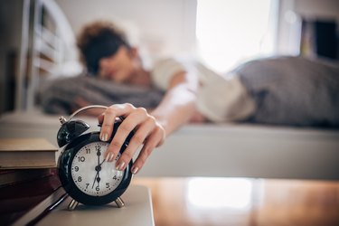 Close up of a person wearing a black sleep mask and white pajamas reaching out of bed to turn off their black alarm clock on their wooden side table