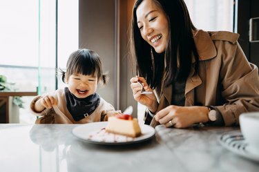 a parent with long brown hair and a young toddler with pigtails sitting at a table sharing a slice of cake