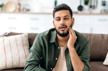 a person with dark hair and beard sitting on the couch and holding their face because they have a popping jaw