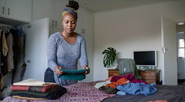 a person with short curly blonde hair wearing a long-sleeved grey shirt and folding laundry on a bed