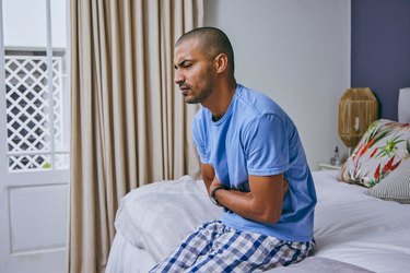 Close up of a person sitting on their bed holding their stomach visibly uncomfortable