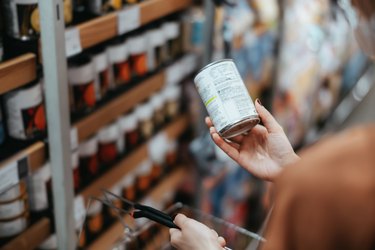 Over the shoulder view of a person reading the nutrition label of a can of food carrying a shopping basket