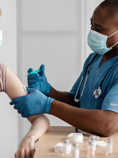 A young black male nurse wearing a mask and giving a shot in the arm to a patient