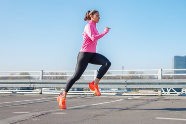 Young fit person with a ponytail wearing a pink jacket, black leggings and orange shoes does a tempo run outdoors