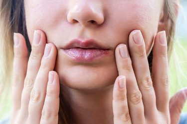 a close up of a person's lips with their hands on their face trying to determine if a red spot is a cold sore vs. a pimple