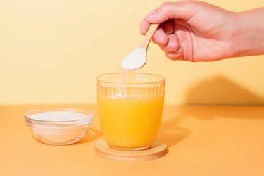 Woman adding collagen powder to orange juice on colorful background