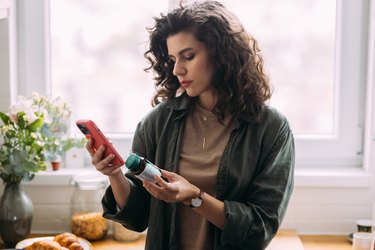 a young adult holds a multivitamin bottle and looks up ingredients on a smartphone with a red case