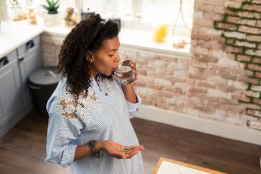 Pregnant person in their kitchen taking prenatal vitamins with a glass of water