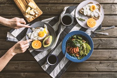 Hand forking into fried eggs (one of the foods that give you energy) on toast with salad and coffee on wooden table