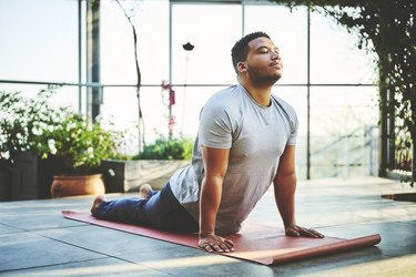 Young man practicing Upward Facing Dog Pose on red mat surrounded by house plants