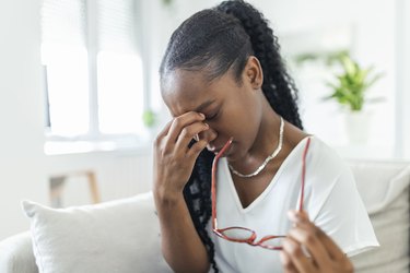 woman at home holding her glasses in one hand and rubbing her itchy eyes with her other hand